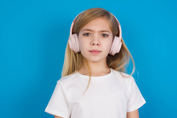 Serious displeased  beautiful caucasian girl wearing t-shirt  looks puzzled at camera being angry wears stereo headphones listens music while walking at street - Photo, Image