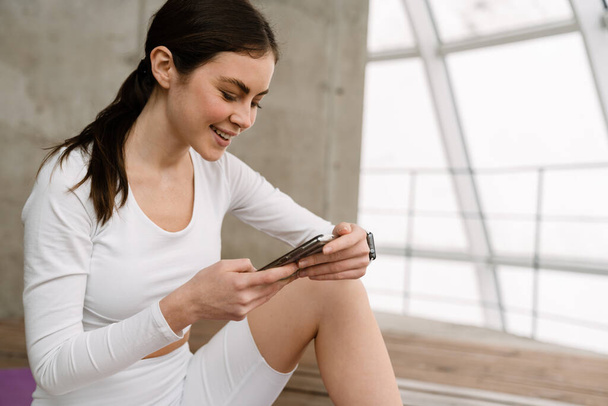 Brunette smiling woman using mobile phone during yoga practice indoors - Photo, image