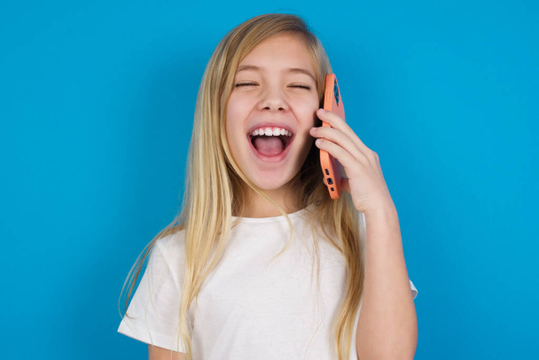 Υπερβολικά χαρούμενη, όμορφη λευκή κοπέλα που φοράει μπλουζάκι γελάει θετικά ακούει αστεία ιστορία από φίλο κατά τη διάρκεια τηλεφωνικής συνομιλίας. - Φωτογραφία, εικόνα