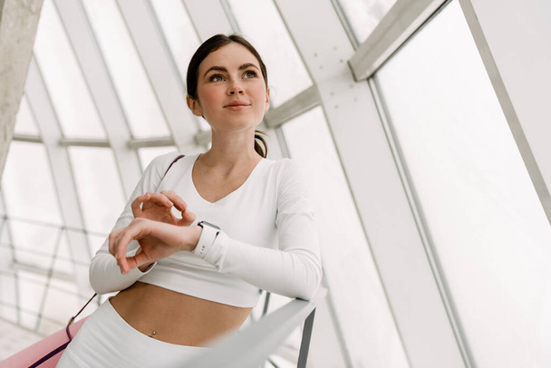 Young pleased woman using smartwatch while standing with yoga mat indoors - Valokuva, kuva