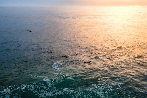 Αεροφωτογραφία των ανθρώπων που περιμένουν τα κύματα για να σερφάρετε στο ηλιοβασίλεμα στον Ατλαντικό Ωκεανό ανοικτά των ακτών Costa da Caparica, Setubal, Πορτογαλία. - Φωτογραφία, εικόνα