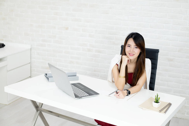 Portrait de belle femme d'affaires asiatique assise derrière la table et reposer le menton sur la main avec le visage souriant dans le lieu de travail.RAF - Photo, image
