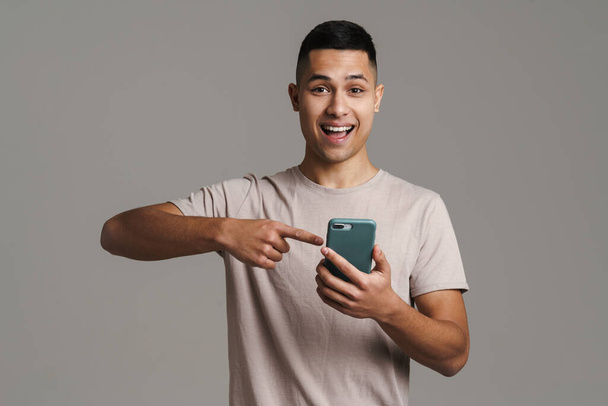 Brunette young excited man holding and pointing finger at mobile phone isolated over grey wall - Zdjęcie, obraz