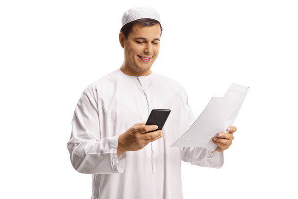 Jonge moslim man in witte kleren met behulp van een smartphone en met een papieren document geïsoleerd op witte achtergrond - Foto, afbeelding