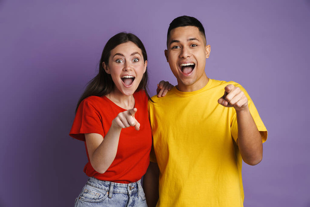 Excited beautiful couple screaming and pointing fingers at camera isolated over violet background - Foto, Imagen