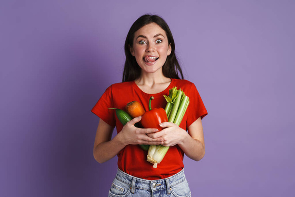 Brunette excited woman showing her tongue while posing with vegetables isolated over purple wall - Fotó, kép