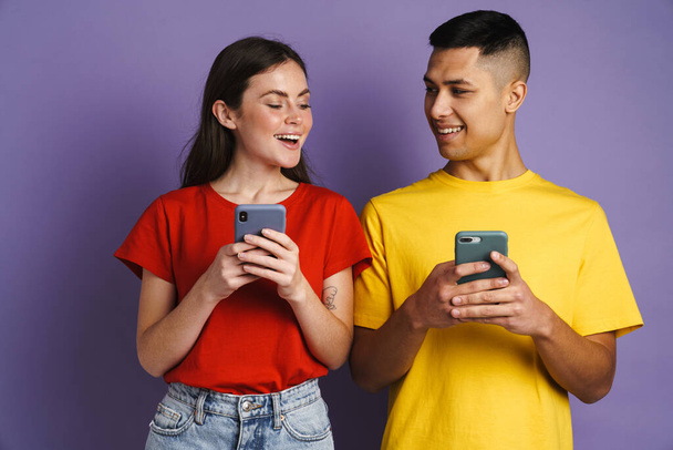 Happy beautiful couple smiling while using mobile phones isolated over violet background - Foto, Imagem