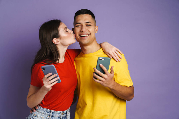 幸せな美しいですカップル接吻しながら使用しています携帯電話絶縁以上紫の背景 - 写真・画像