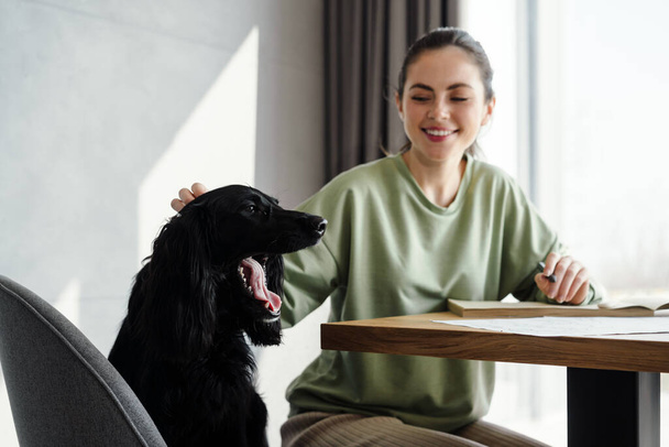Šťastná brunetka mladá žena mazlit svého psa, zatímco studuje doma, sedí u stolu, dělat si poznámky - Fotografie, Obrázek