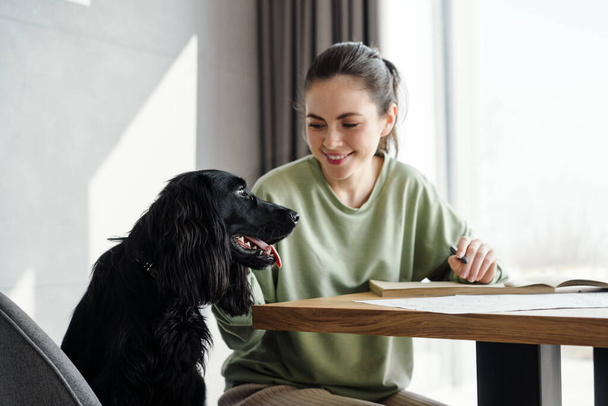 幸せなブルネット若いです女性petting彼女の犬ながら家で勉強,座ってテーブル,ノートを取ります - 写真・画像