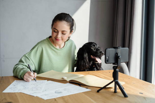 Happy young brunette woman studying online remotely from her house while sitting in front of a mobile phone during video call, petting dog - Fotó, kép