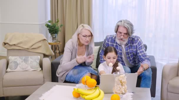 Isovanhemmat katselee kannettavaa tietokonetta ja puhuu samalla auttaa pieni tyttö lapsenlapsi koulun kotitehtäviä - Materiaali, video