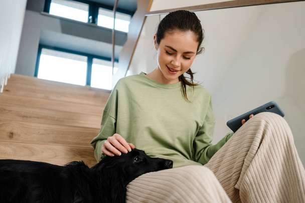 Smiling young brunette woman petting her dog at home, sitting on steps, holding mobile phone - Foto, Imagem