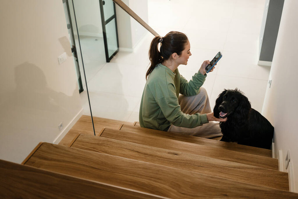 Smiling young brunette woman petting her dog at home, sitting on steps, holding mobile phone - Photo, image