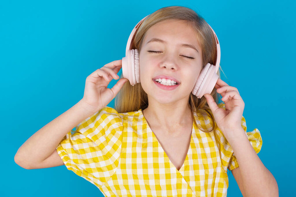 schöne kaukasische Mädchen im Kleid lächelt breit fühlt sich sehr froh hört Lieblingsmusik über drahtlose Kopfhörer schließt die Augen. - Foto, Bild