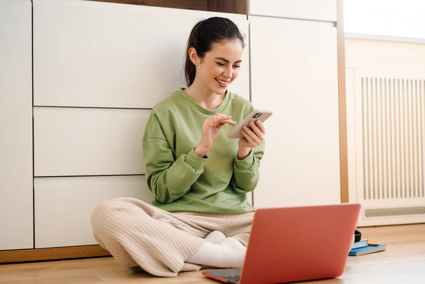 Smiling young brunette woman using mobile phone while sitting with laptop computer on a floor at home - Photo, Image