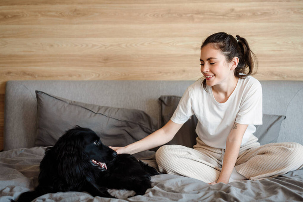 Happy young brunette woman with her dog at home sitting on a sofa - Fotó, kép