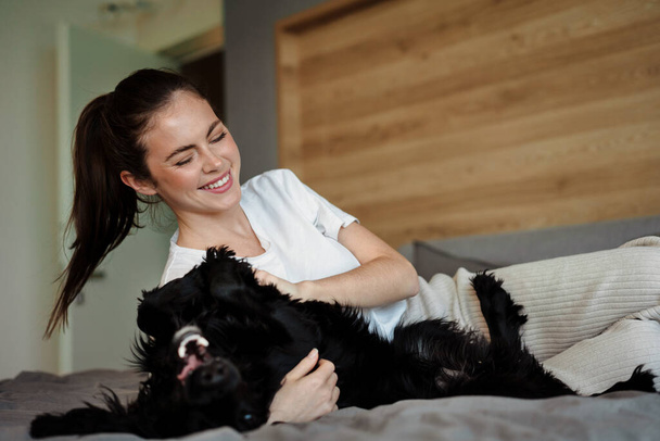 Smiling young brunette woman petting her dog at home, laying on a couch - Φωτογραφία, εικόνα