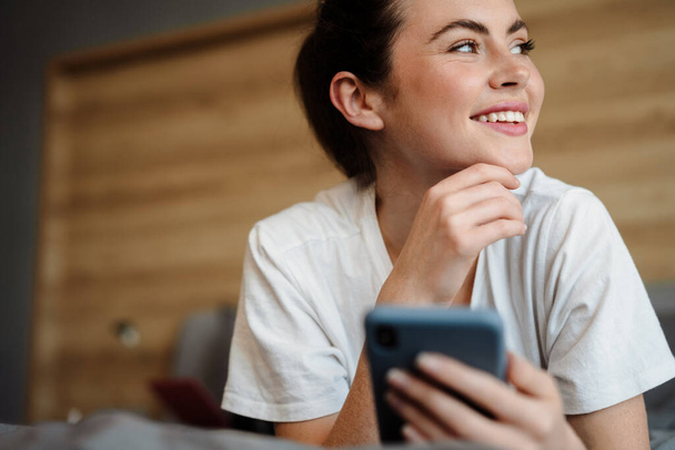 Sorridente giovane donna bruna che tiene il telefono cellulare sdraiato sul letto in casa, distogliendo lo sguardo, da vicino - Foto, immagini