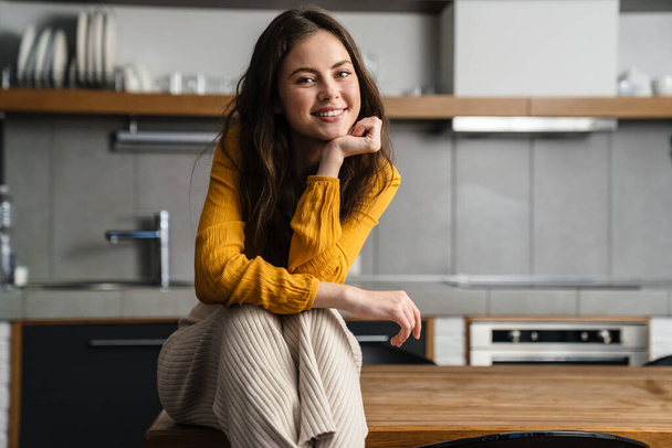 Young brunette woman smiling at camera while sitting on table in home kitchen - Foto, immagini