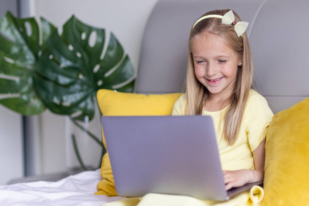 Cute little caucasian girl with blonde hair in fashionable dress illuminating yellow color sitting at home during coronavirus pandemic quarantine and using laptop. Stay at home during covid-19 - Photo, Image