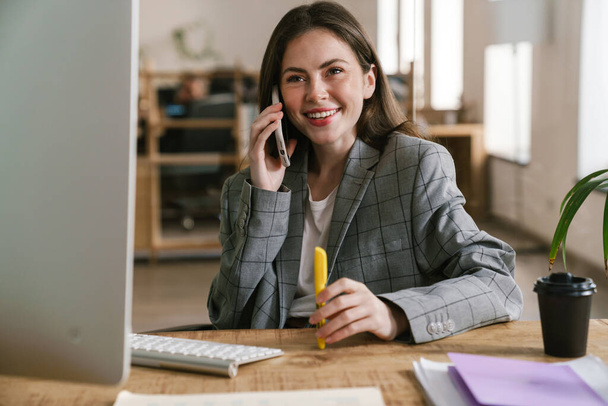 Smiling brunette woman talking on mobile phone while working with computer in office - Photo, Image