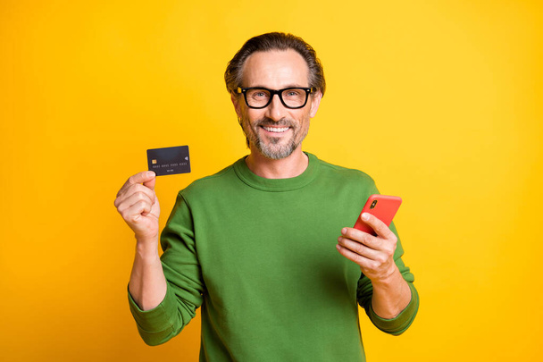 Photo of cheerful man demonstrate mobile phone debit card wear spectacles green pullover isolated yellow color background - Foto, Imagen