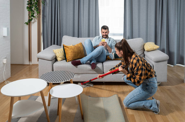 Mujer joven limpiando el piso del apartamento mientras el hombre perezoso sentado en el sofá y jugando juegos en su teléfono inteligente - Foto, Imagen