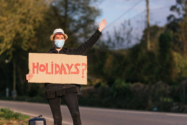 Şapkalı ve antivirüs maskeli genç bir kız tabelasıyla yolda durup tatilleri soruyor. Tatil kaçamağı. İstikamet yok. genç kültür - Fotoğraf, Görsel