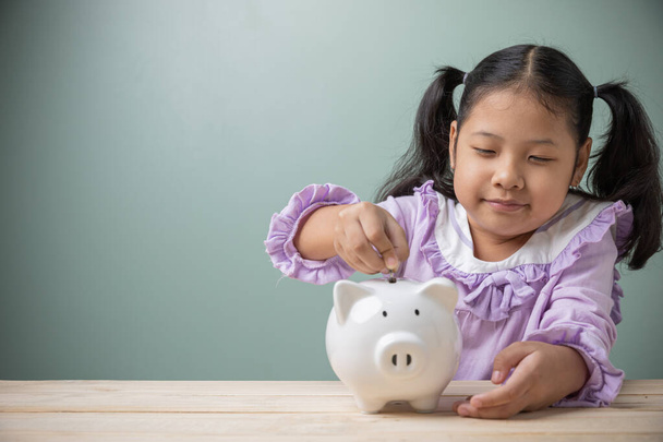 An Asian kid girl put a coin in a white pig piggy bank on a wood table at home. Save money for tuition fees, the cost of school supplies in the future. Ideas to save money for education future use. - Foto, Bild