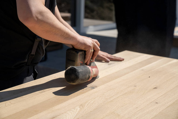 A construction worker using a cordless power sander to sand a massive wooden table top - Photo, Image