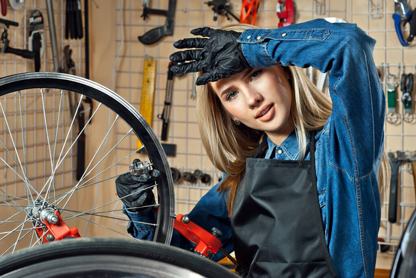 Woman in a bicycle workshop on the background of tools. Blonde in a blue denim shirt repairs a bicycle. The concept of professional bicycle service. - Foto, afbeelding