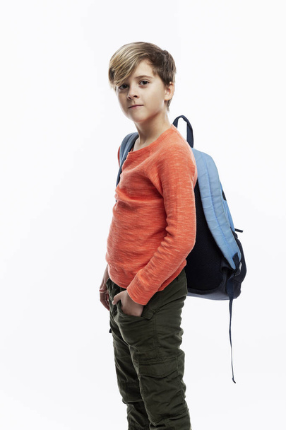 A 9-year-old boy in an orange jacket and jeans stands holding his hands in his pockets with a school backpack on his shoulder. White background. Vertical. - 写真・画像