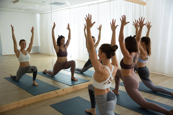 Group of young sporty women in yoga studio, practicing yoga pose in front of the mirror. Healthy active lifestyle, working out indoors in gym - Photo, Image