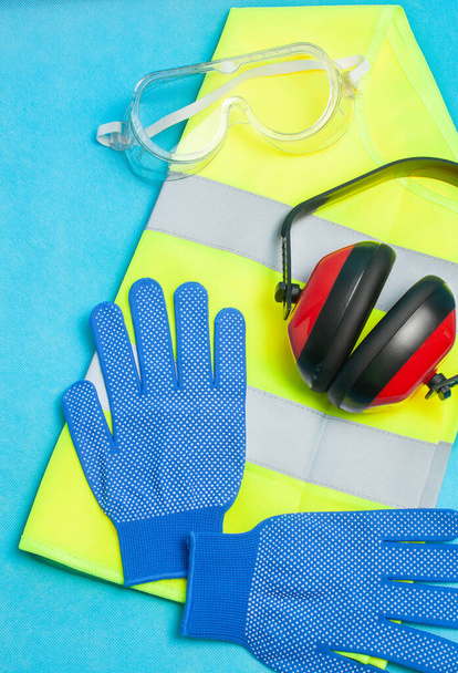 Vista de vários elementos de proteção para a segurança no trabalho, como óculos, colete reflexivo, capacetes de proteção auditiva e luvas em um fundo azul . - Foto, Imagem