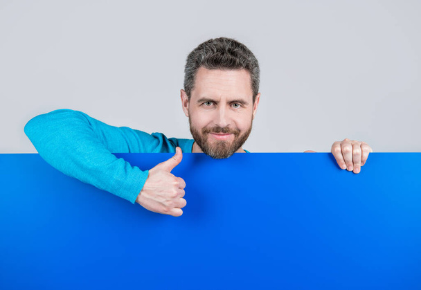 fröhliche reife bärtige Mann hinter leeren blauen Papierbanner mit Kopierraum für Präsentation zeigen Daumen hoch, Kommunikation. - Foto, Bild