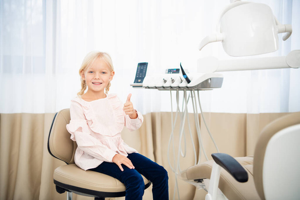 Крупним планом маленька дівчинка в стоматологічному кабінеті показує великі пальці вгору. Діти відвідують концепцію стоматологів. Концепція охорони зубів
 - Фото, зображення