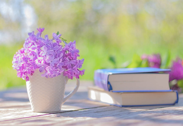 pequeño ramo de lilas florecientes en una taza blanca y libros cerrados sobre la mesa en el jardín - Foto, Imagen