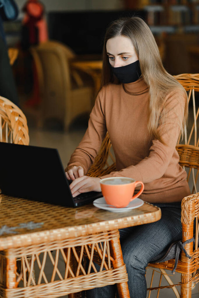 young girl in a black medical mask work in cafe on a laptop - Photo, Image