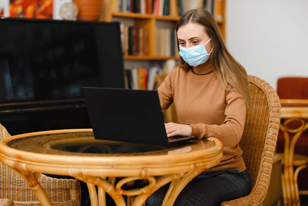 young girl in a blue medical mask work in cafe on a laptop - Foto, imagen