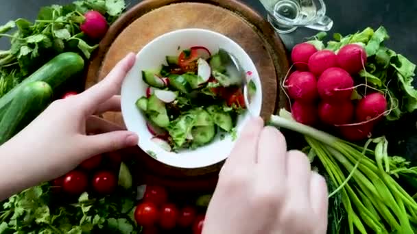 Mani mescolare l'insalata di verdure con un cucchiaio. Intorno alla ciotola una pila di verdure fresche e mature: ravanelli, pomodori, cetrioli, erbe aromatiche e aglio. - Filmati, video