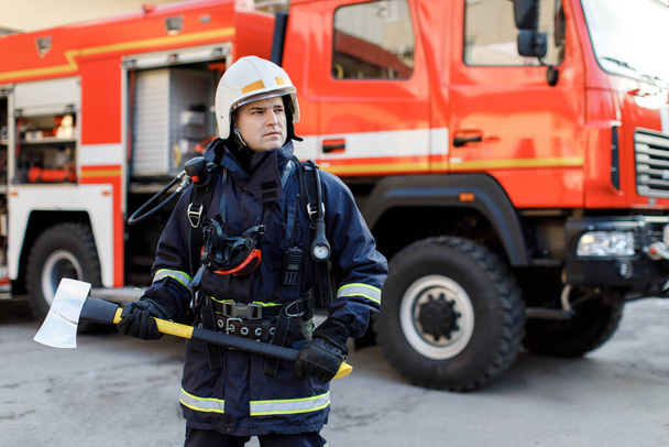 Portret poważnego i pewnego siebie białego strażaka stojącego i trzymającego młotek, noszącego specjalny mundur ochronny w tle ciężarówki. - Zdjęcie, obraz