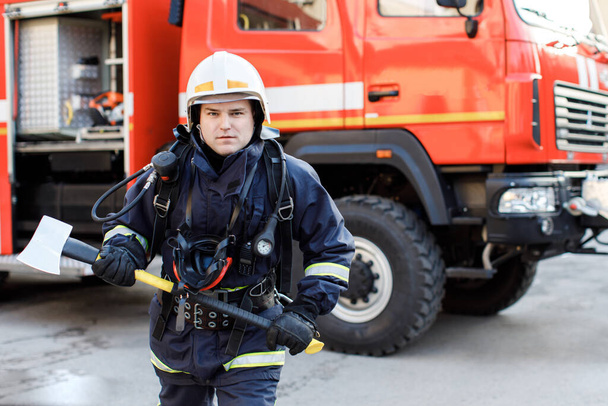 Vakavan ja itsevarman valkoihoisen palomiehen muotokuva seisomassa ja pitämässä vasaraa, yllään erityinen suojaava univormu kuorma-auton taustalla. - Valokuva, kuva