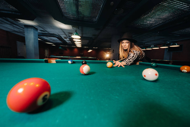 A girl in a hat in a billiard club with a cue in her hands hits a ball.Playing pool. - Fotografie, Obrázek