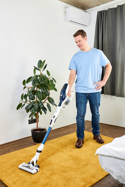 Handsome caucasian man cleaning floor carpet with vacuum cleaner in modern white living room - Photo, Image