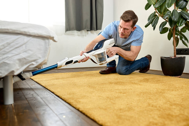 В современной белой гостиной мужчина моет пол с помощью вакуумной чистки. Концепция легкой очистки с современным новым пылесосом - Фото, изображение
