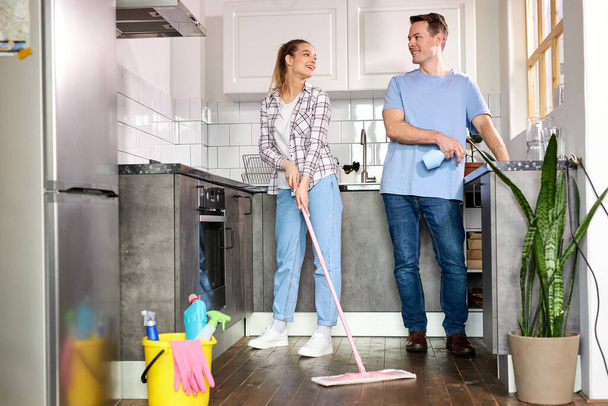 Friendly Young Couple Cleaning Home, In Kitchen. Woman Wiping Floor With Mop, Husband Helping - 写真・画像