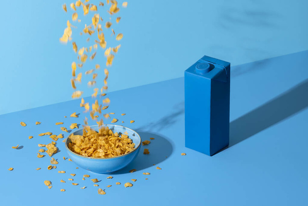 Granen in een kom gieten op een blauw gekleurde tafel. Ontbijttafel met cornflakes ontbijtgranen in een kom en een melkbox, bij helder licht. - Foto, afbeelding