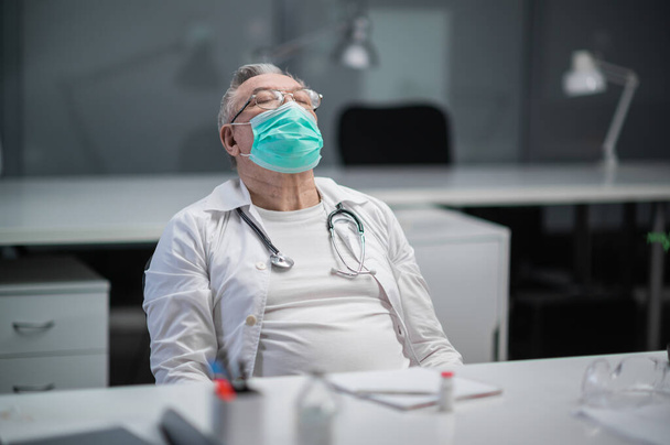 Літній досвідчений лікар у медичній масці нахилився назад у свій кабінет, він був дуже втомлений після робочого дня під час епідемії
 - Фото, зображення