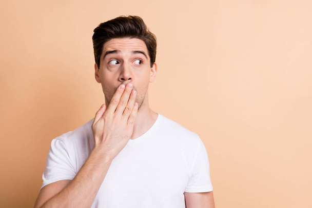 Photo of astonished young guy cover hand mouth look empty space isolated on pastel beige color background - 写真・画像